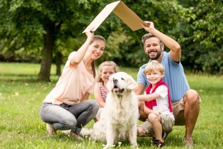 Duurzaam wonen: gezin beschermt huis en hond met kartonnen dak in groene tuin, symbool voor woningbescherming.