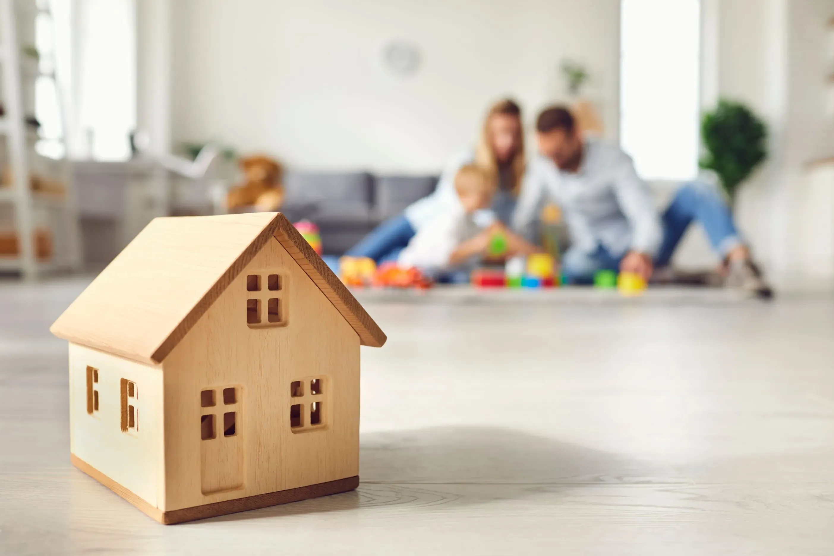 Gezin speelt in de woonkamer op de achtergrond. Op de voorgrond staat een houten huisje. 